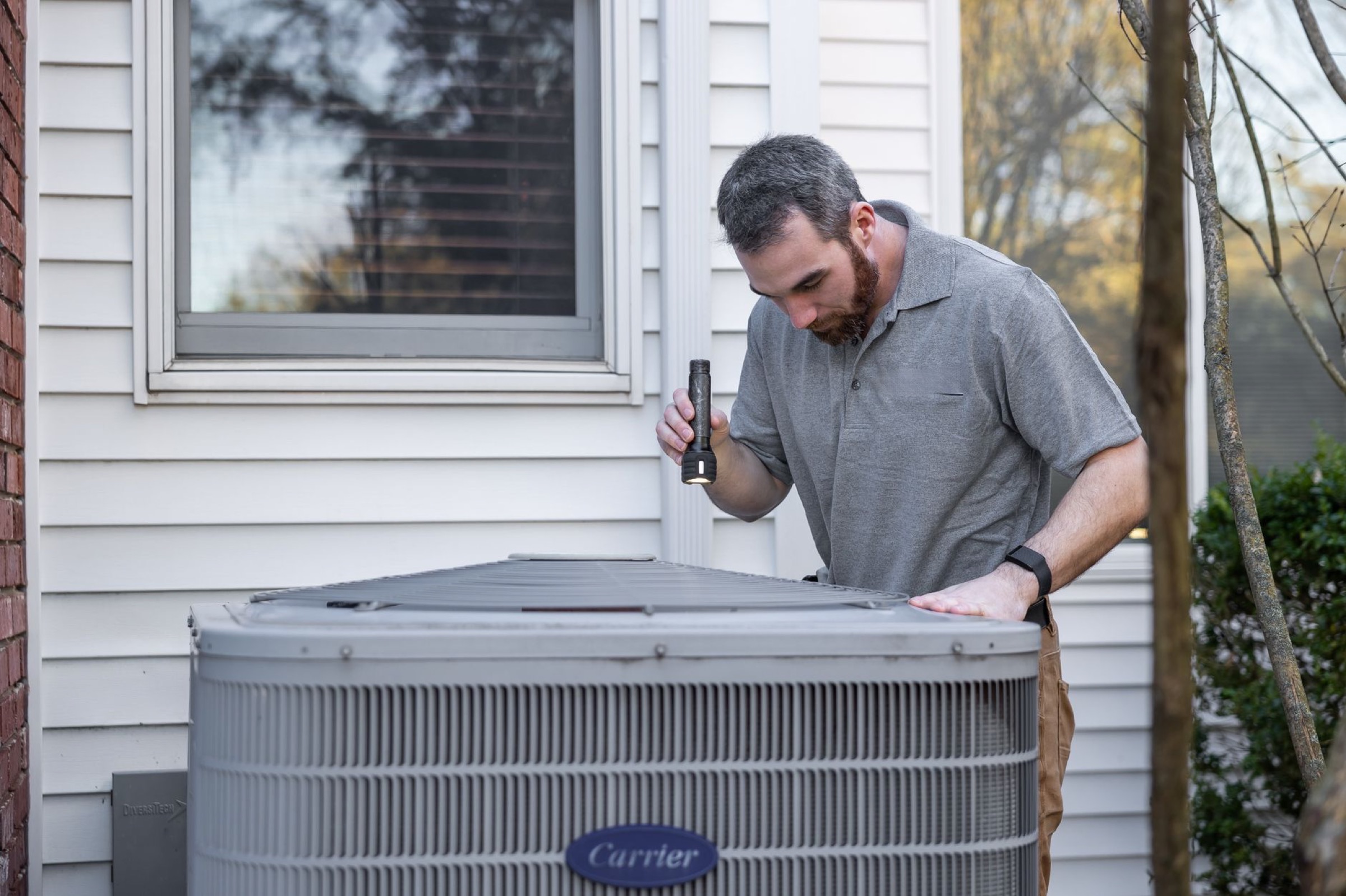 An HVAC technician inspecting an outdoor condenser with a flashlight