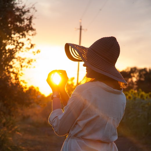 A woman in a wide hat holding the setting sun in her hand