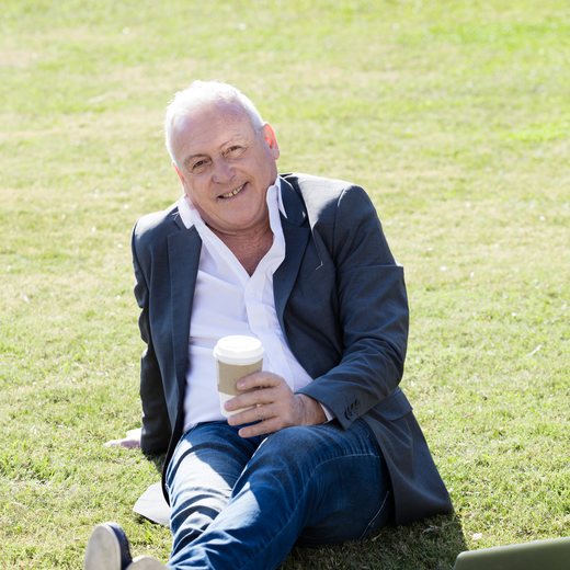 A silver-haired man in a blazer sitting on the grass with a coffee, laptop beside him
