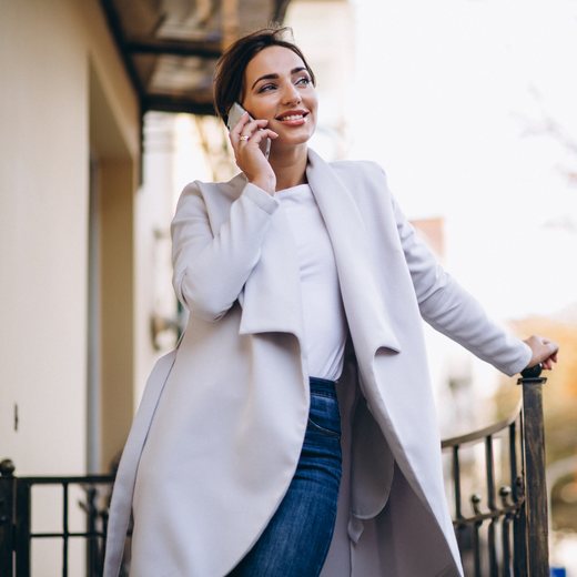 A woman in a pale coat taking a call outdoors in soft daylight