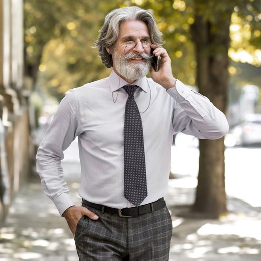 An older man in shirt and tie on the phone, walking a tree-lined street