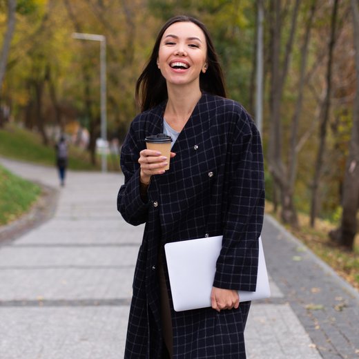 A woman laughing on a park path, carrying a laptop and a coffee